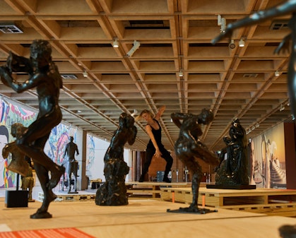 In a large gallery space, a person dressed in black moves, surrounded by black figure sculptures.