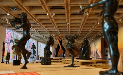 In a large gallery space, a person dressed in black moves, surrounded by black figure sculptures.