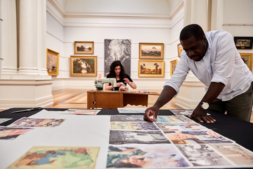 One person leans over a table which holds a series of images. Another person works at a sewing machine.