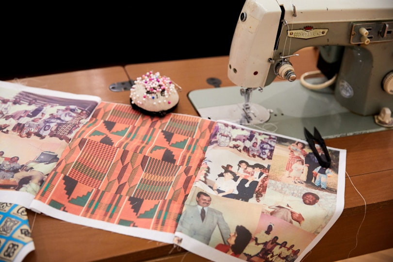 Material printed with a series of photos and a graphic design sits on a table beside a sewing machine and a pincushion.