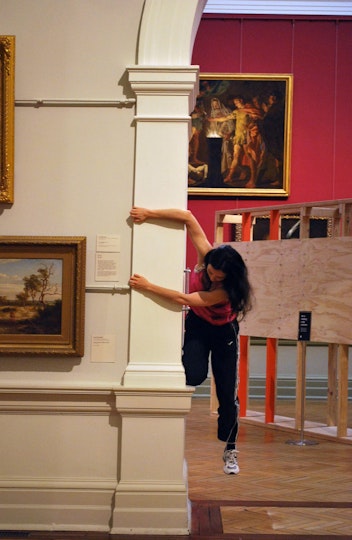 A person kneels with one leg on a small ledge in a large archway between two rooms in a gallery. They hold the wall with both hands as their other leg dangles.