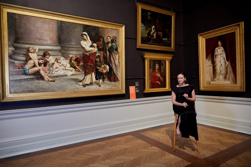 A person sits on a stool in a corner of a gallery between gold-framed historic paintings.
