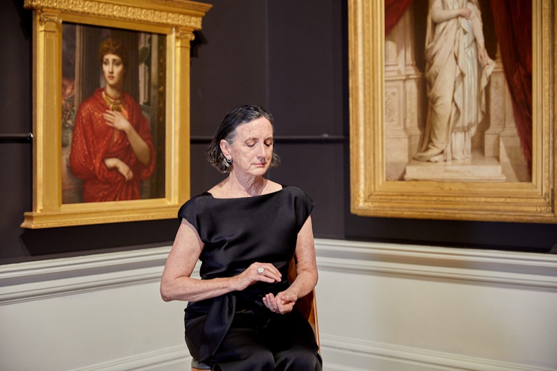 A person sits in a corner of a gallery between gold-framed historic paintings, with their eyes closed and their hands cupped one above the other.