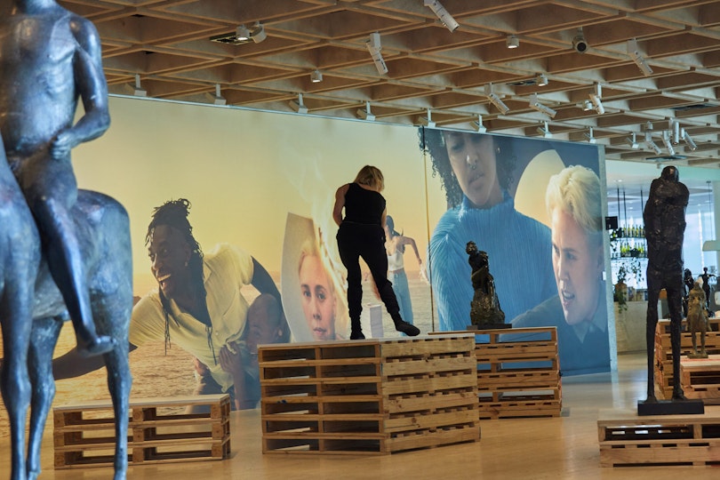 In a gallery space, a person stands in a slightly twisted position on top of a packing crate. Around them are three figure sculptures and a mural depicting several people. 