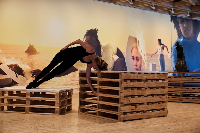 A person balances sideways between two packing crates of different heights. Behind them is a figure sculpture and a mural depicting people and water.