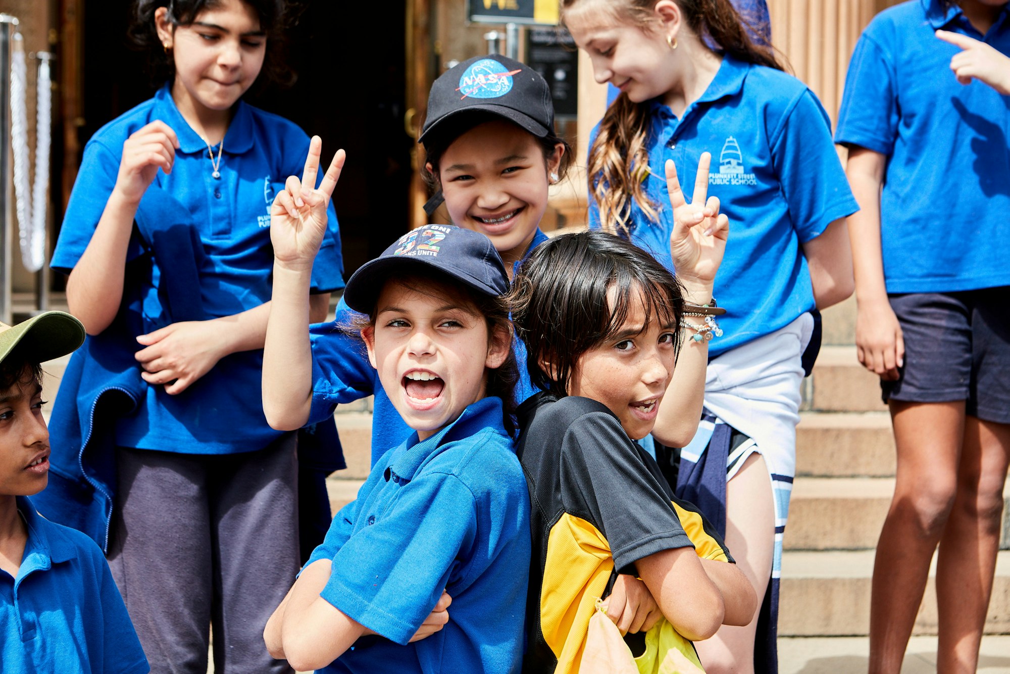 A group of several children, all but one dressed in the same blue collared t-shirt. Two of the kids stand back to back with arms crossed. Another stands behind them making a V sign with each hand.