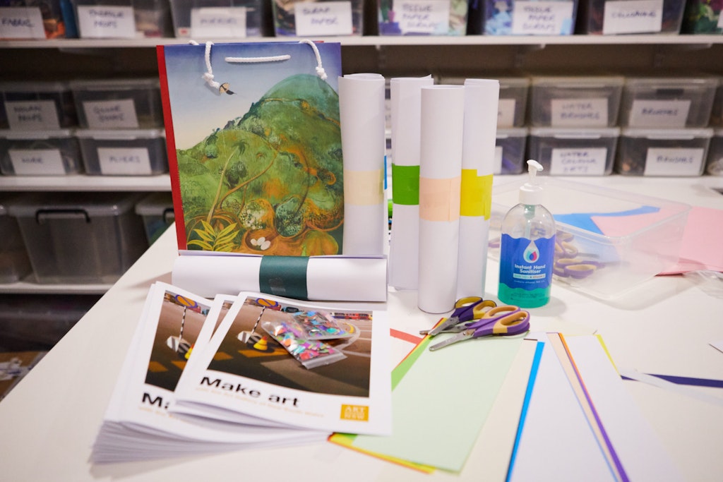 On a table sits a pile of booklets titled 'Make art' as well as paper, scissors, a carry bag and a bottle of hand sanitiser.