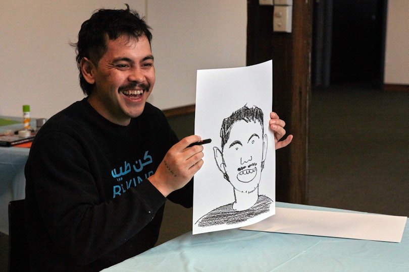 A laughing person with short hair and a moustache sits at a table and holds up a self-portrait drawing.