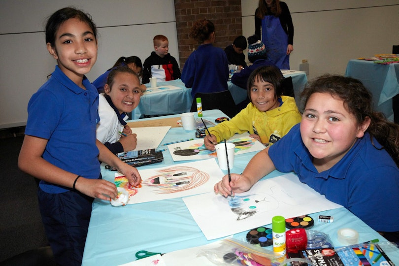 A group of smiling students create atworks on a table on which sit various art supplies.