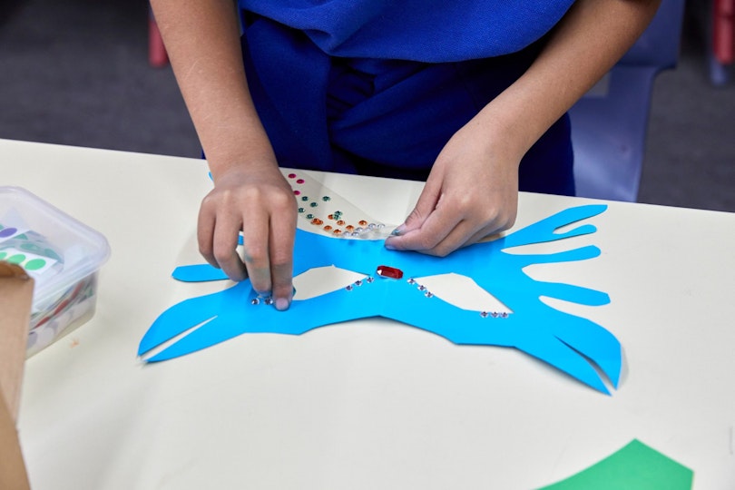 Two hands decorate a blue cardboard mask with small shiny coloured dots.
