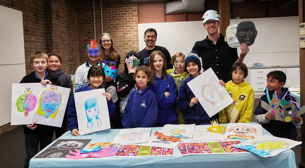 A group of people stand behind a a table covered with artwork images. Some of the people hold up drawings or masks, or are wearing decorative masks.