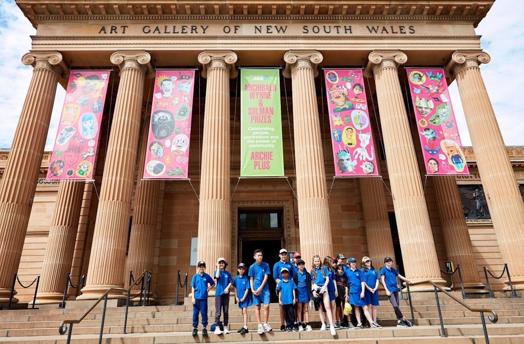 A group of children in blue uniform stand in front of a sandstone building with 'Art Gallery of New South Wales' above its entrance. Five banners hang between columns. Four of these feature multiple illustrations on a pink background.