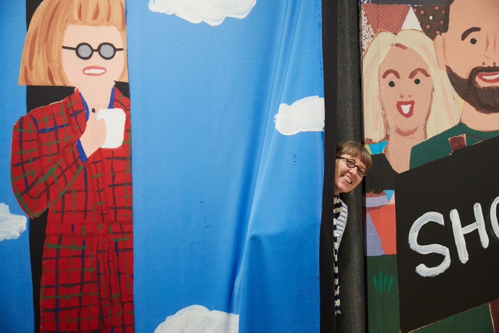 A person with fringed hair and glasses peaks out from a blue curtain with white clouds. There are painted people on either side, included one with fringed hair and glasses.