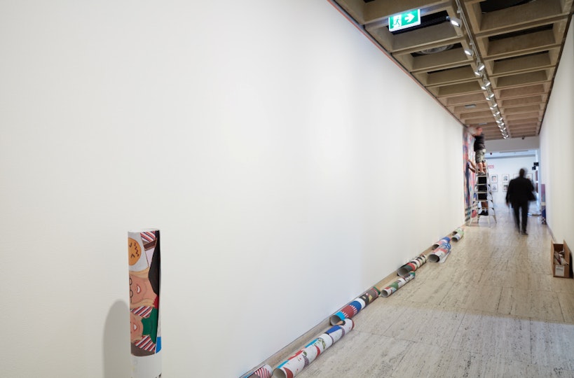 A long white wall with large tubes of rolled-up printed paper in front of it.