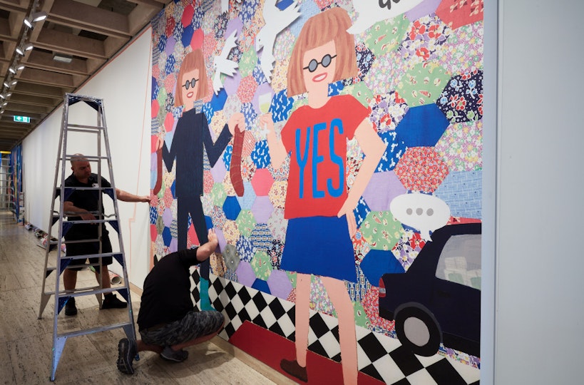 A person kneels in front of a section of artwork on a wall while another person stands next to a ladder.