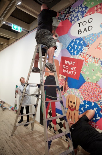 A person with fringed hair and glasses stands on the ground next to a person on a ladder in front an artwork on a wall.
