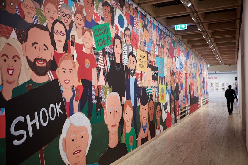 A person walks down a long hallway where there is a colourful mural depicting a multitude of individuals.