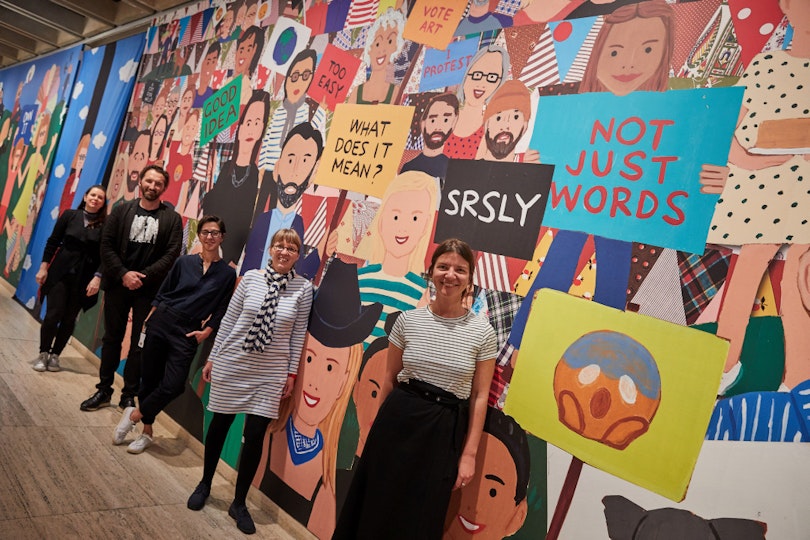 Five people stand in front of a large mural depicting a multitude of individuals, some holding signs.