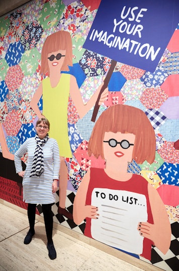 A person with fringed hair and glasses stands next to an artwork depicting two figures who also have fringed hair and glasses. One holds a sign that says 'Use your imagination'. The other holds a piece of paper that is headed 'To do list...'