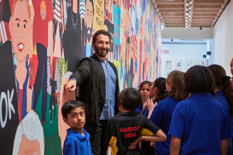A bearded person stands smiling and pointing among a group of children in front of a mural.