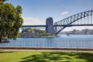 A view over grass and through a low paling fence across water to the Sydney Harbour Bridge