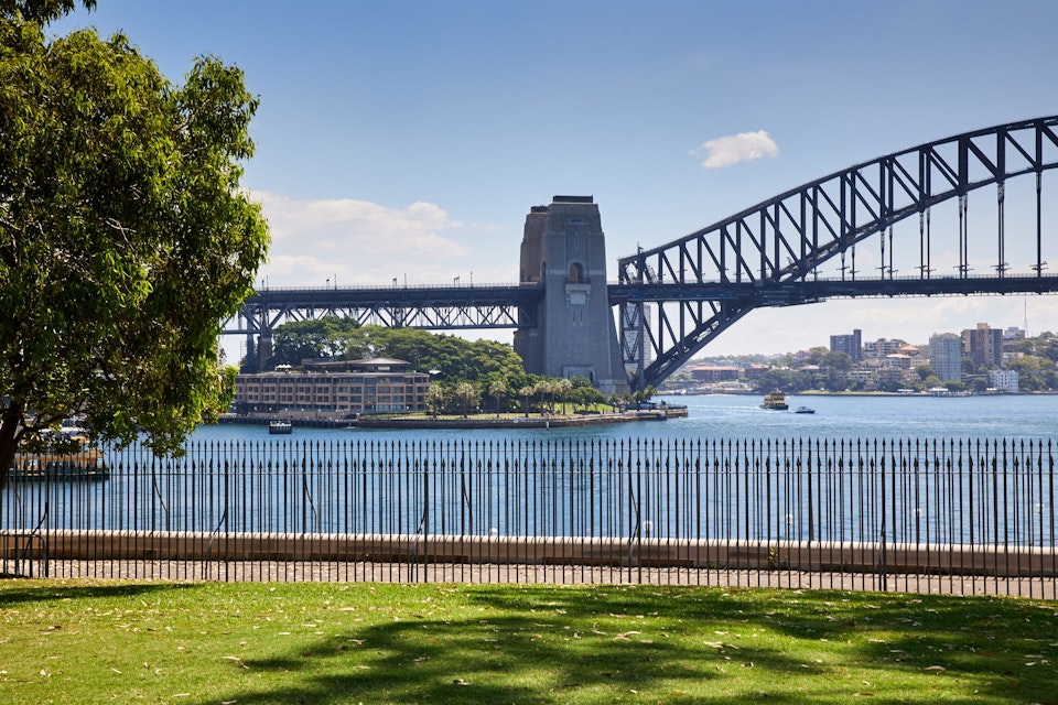 A view over grass and through a low paling fence across water to the Sydney Harbour Bridge