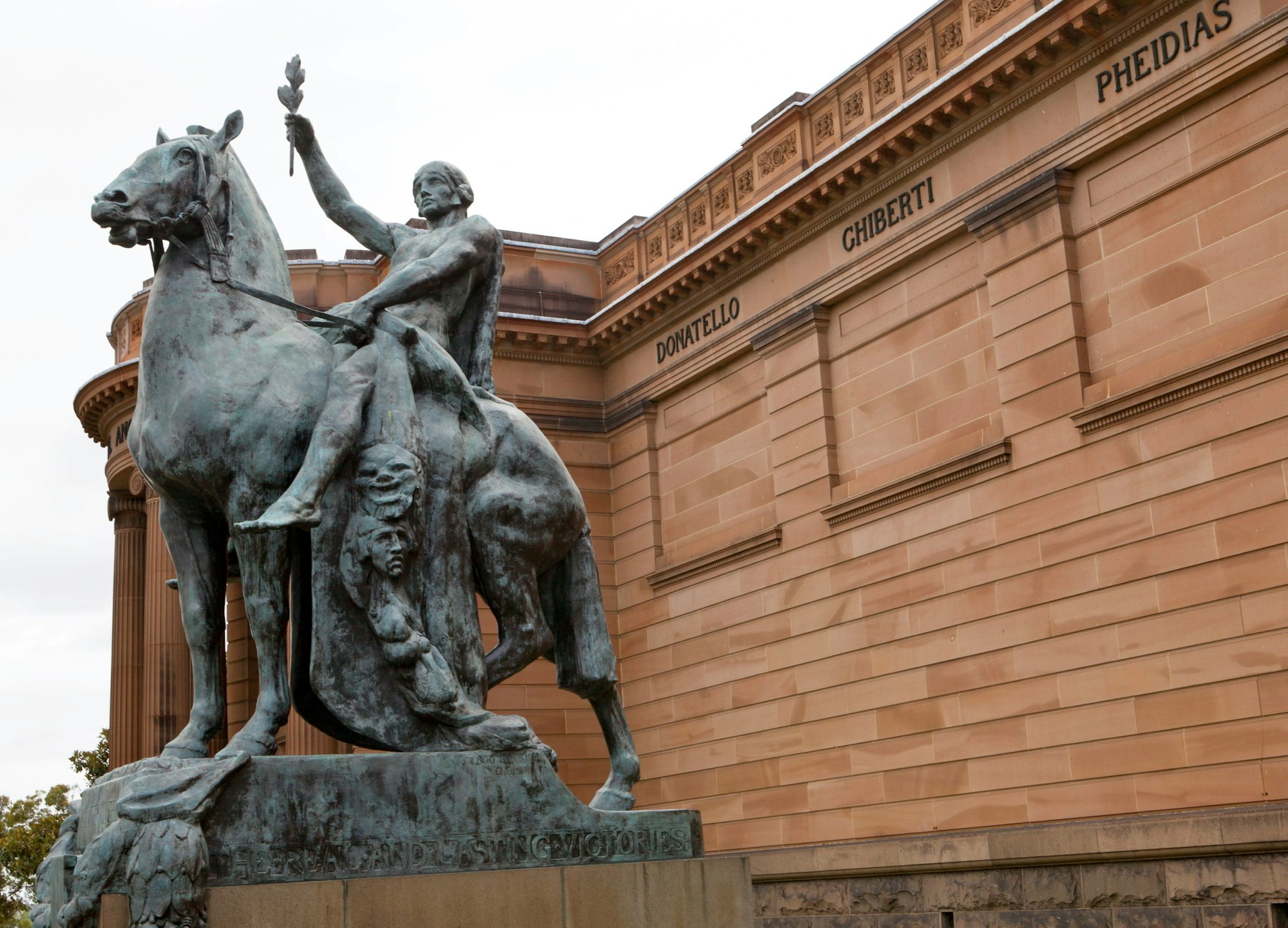 A statue of a person holding up a branch while seated on a horse in front of a large sandstone building.