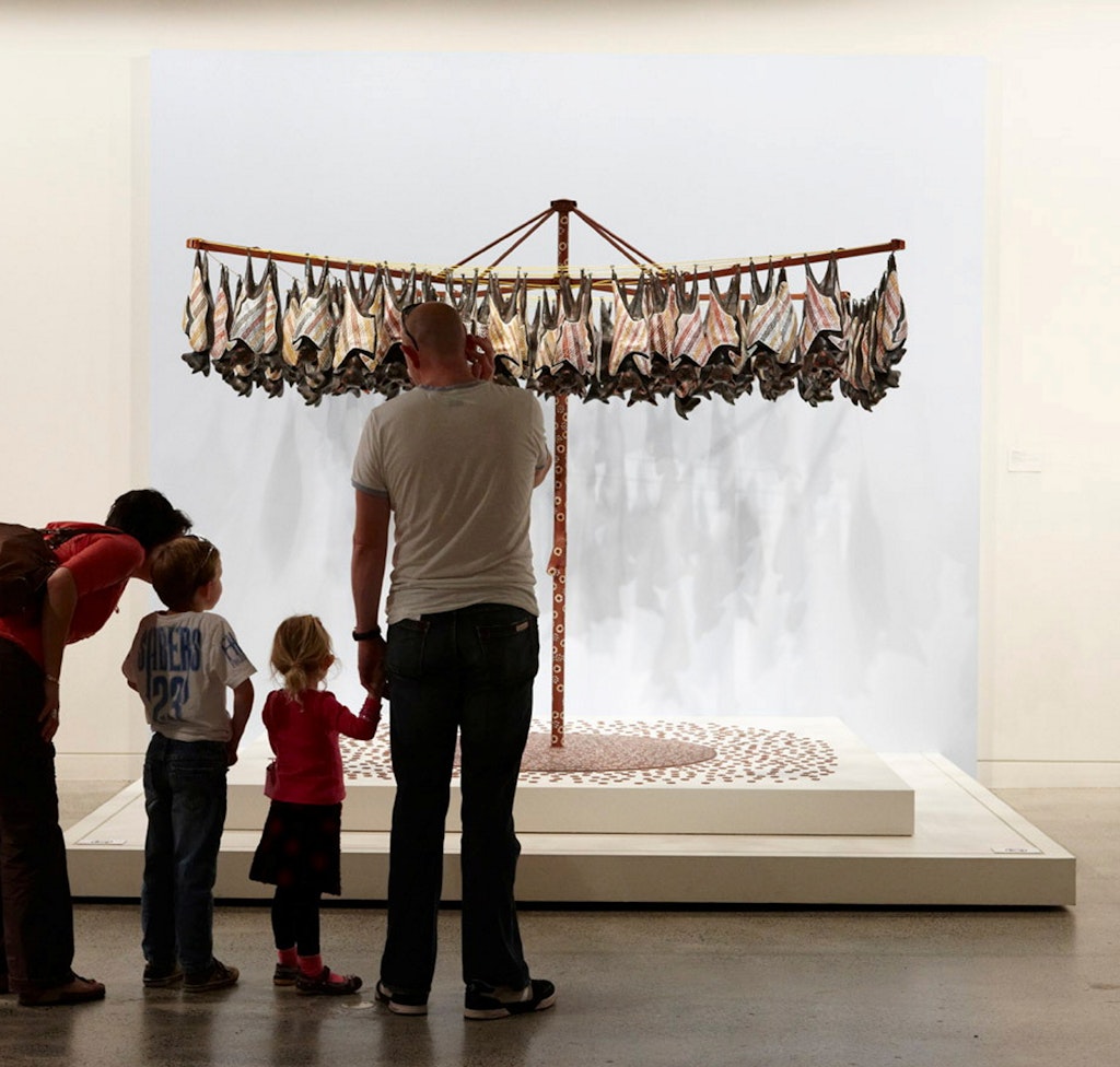 People of different ages stand looking at an artwork of painted fruit bats hanging upside down from a Hills Hoist-like washing line.