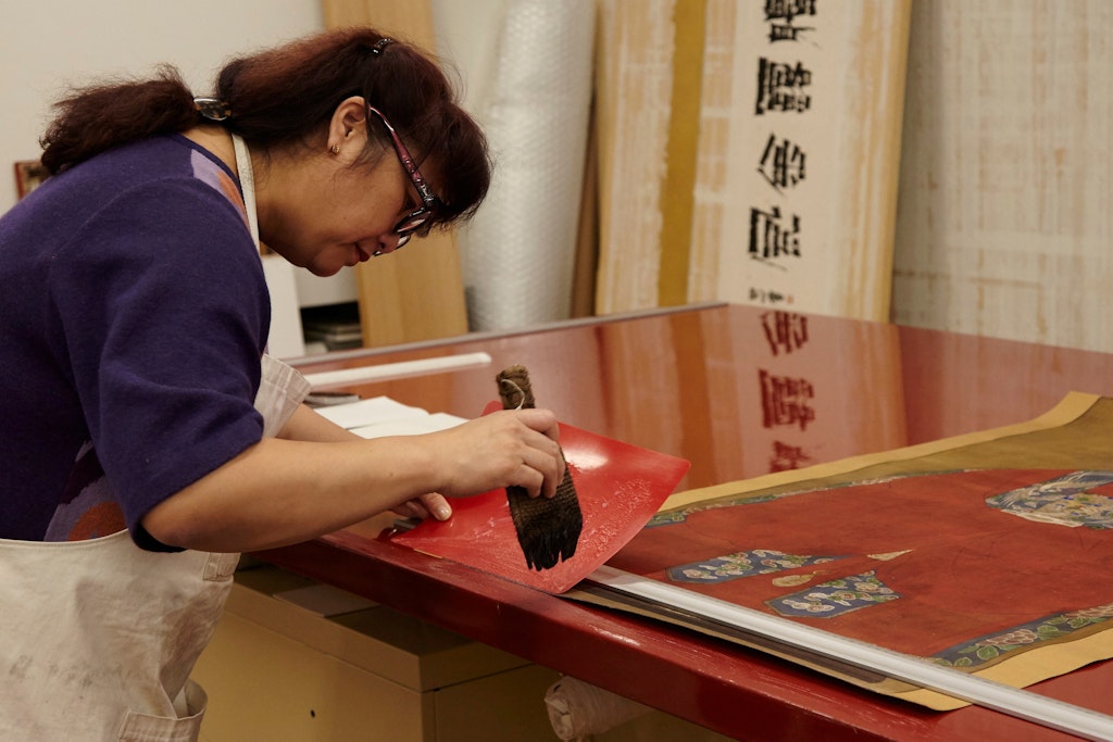 A person holds a wide brush while bending over an artwork lying on a work bench.