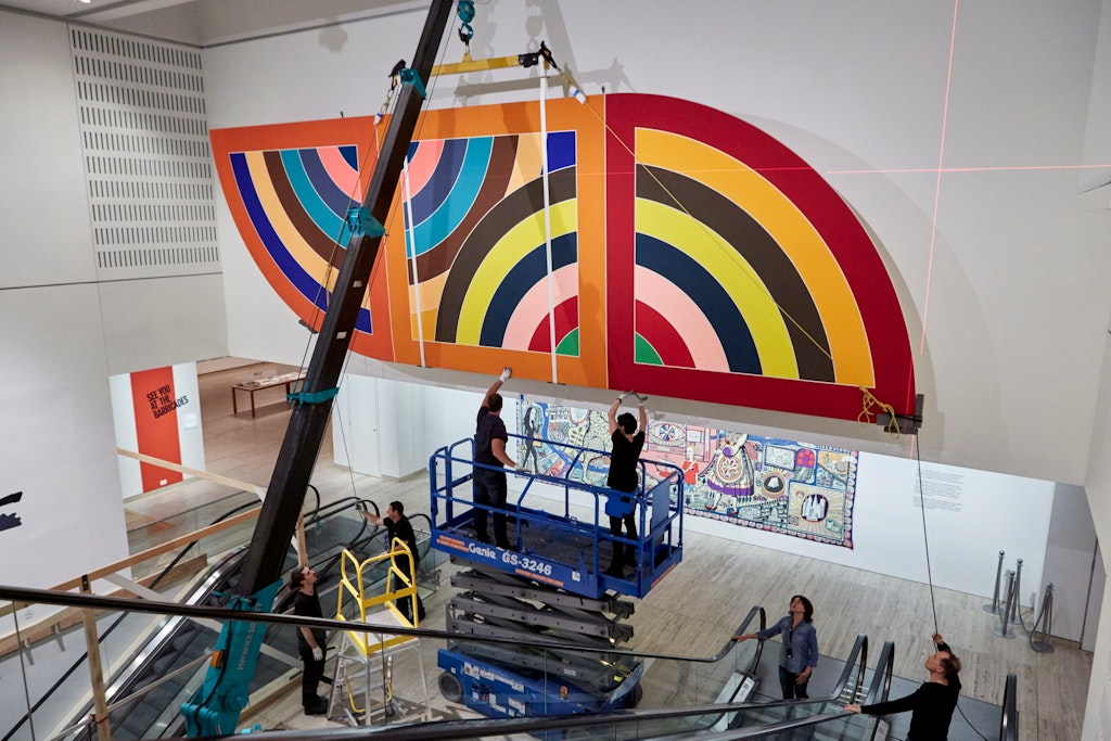 A crane moves a massive abstract painting into place on a high wall assisted by two people on a scissor lift and other people on the ground.