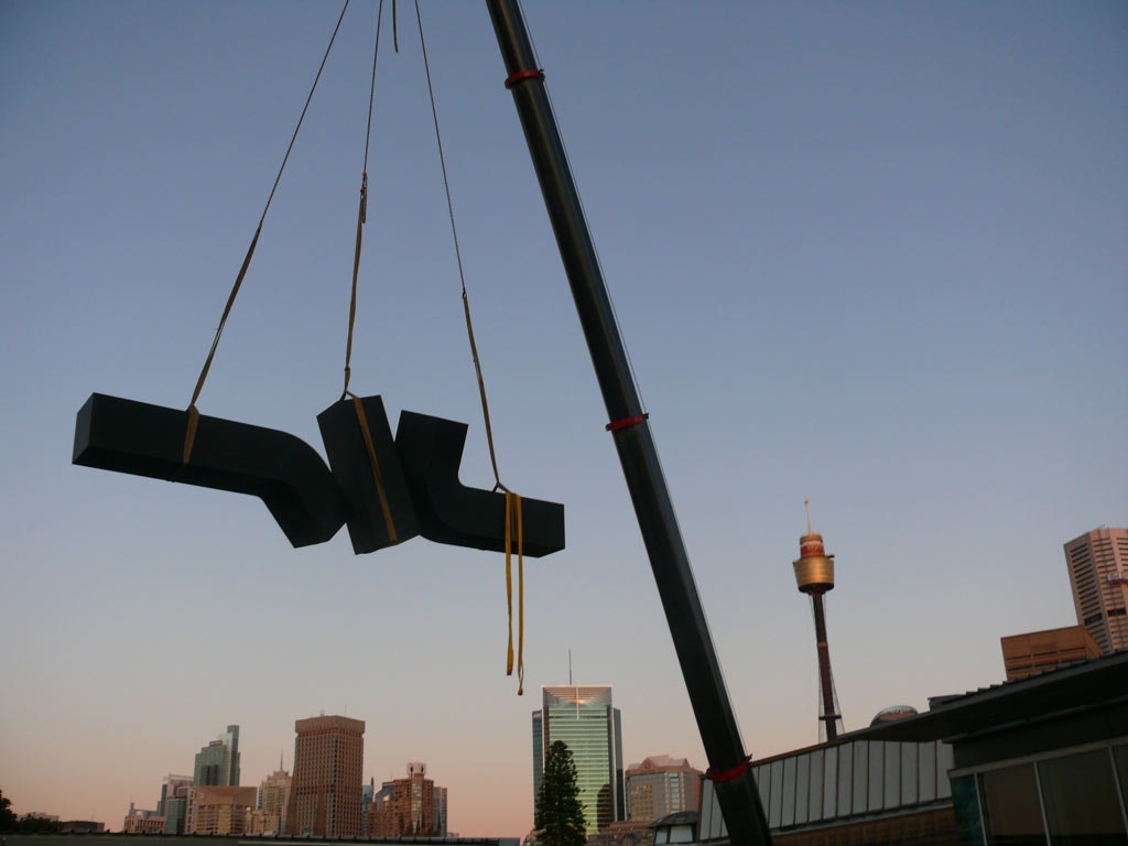 A massive sculpture of three black rectangular shapes is lifted by a crane, with the Sydney skyline in the background.