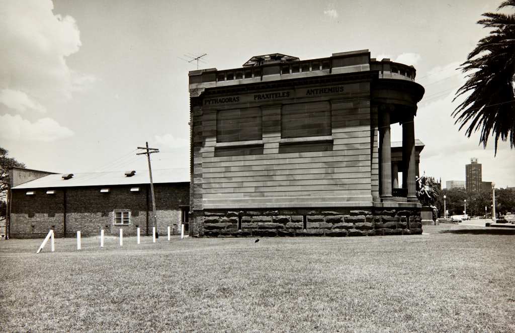Black-and-white photograph of an imposing sandstone oublic building with a small, unimposing building at the back.