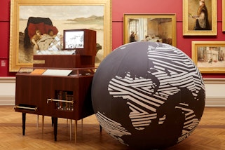 In a red-walled gallery hung with gold-framed paintings a large globe of the world sits on the floor next to a wooden cabinet.