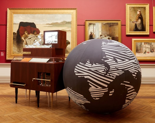 In a red-walled gallery hung with gold-framed paintings a large globe of the world sits on the floor next to a wooden cabinet.