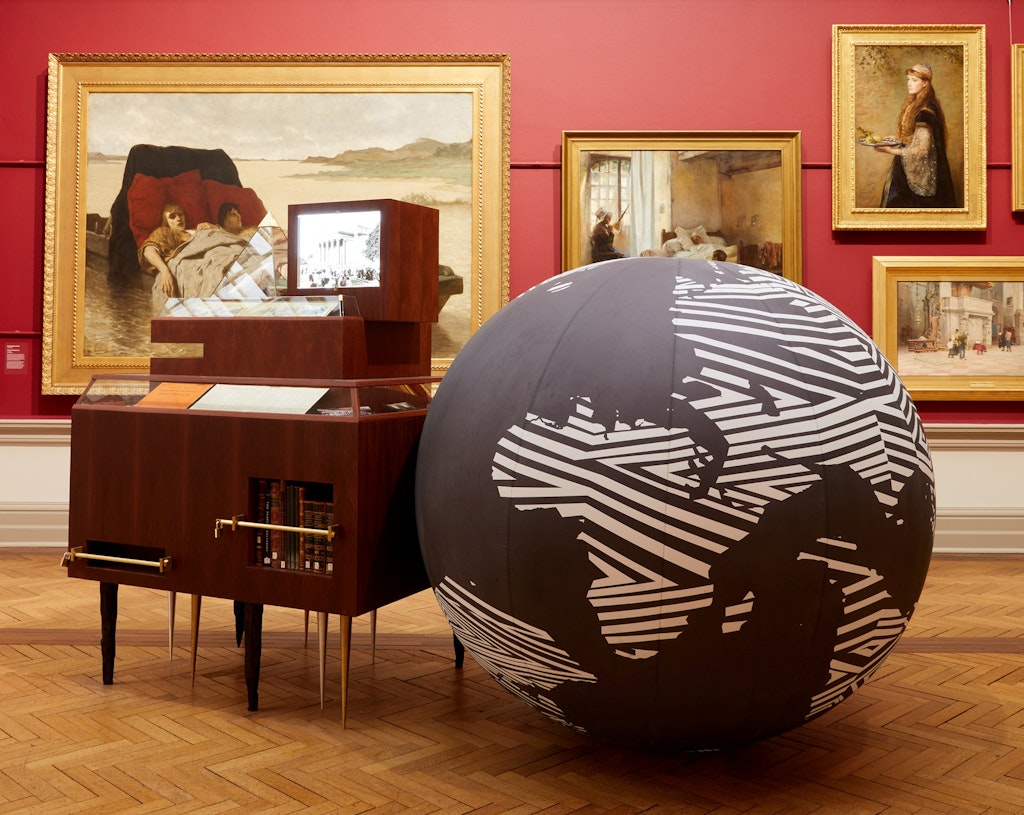 In a red-walled gallery hung with gold-framed paintings a large globe of the world sits on the floor next to a wooden cabinet.