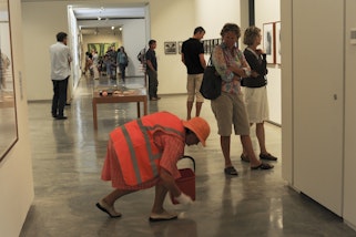 A performance by Julie-Anne Long called 'Val, the Invisible' Image shows a middle aged women in a high visibility vest cleaning the floor of the Gallery with visitors looking on.