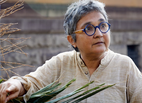 A person with short grey hair and round glasses holds a large palm leaf.