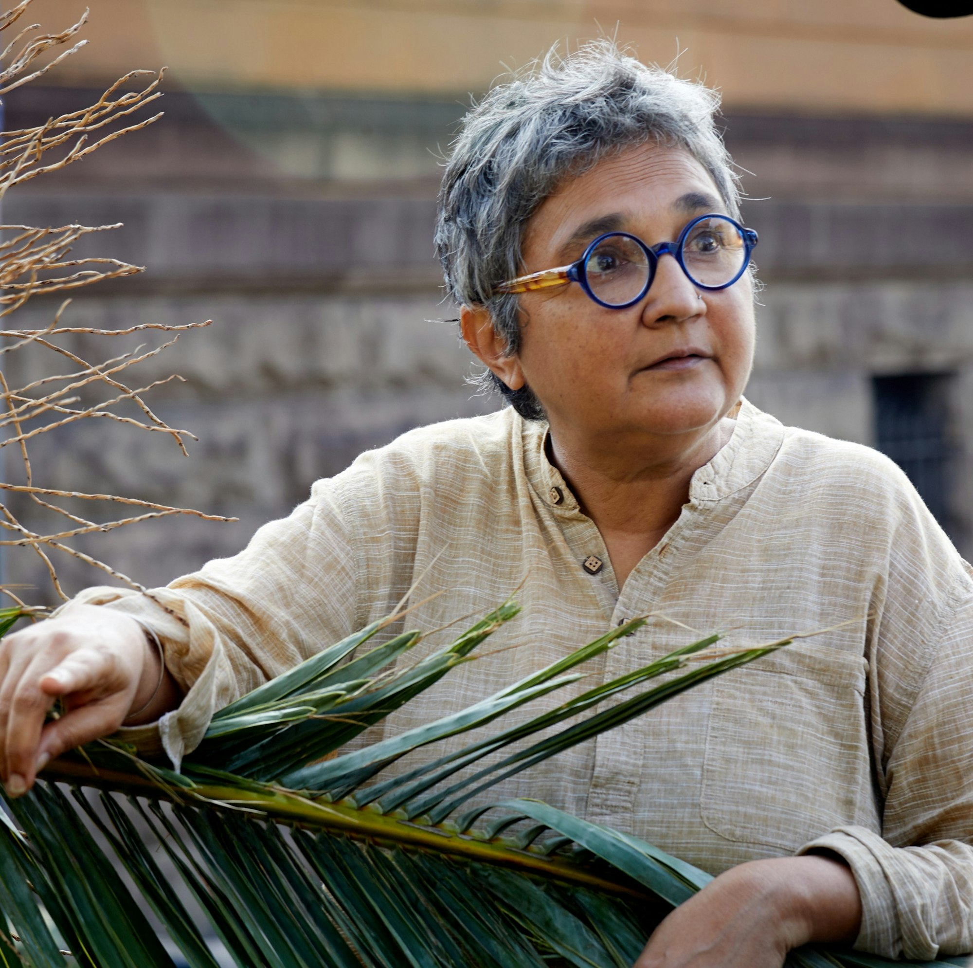 A person with short grey hair and round glasses holds a large palm leaf.