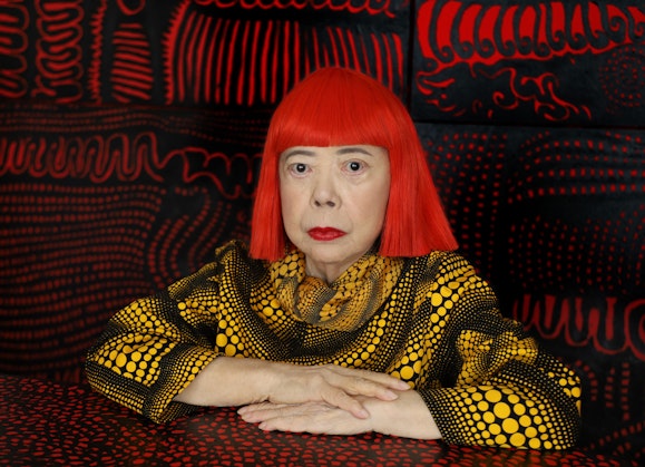 A person with bright red bobbed hair and a yellow-spotted black top  in a red and black patterned space.