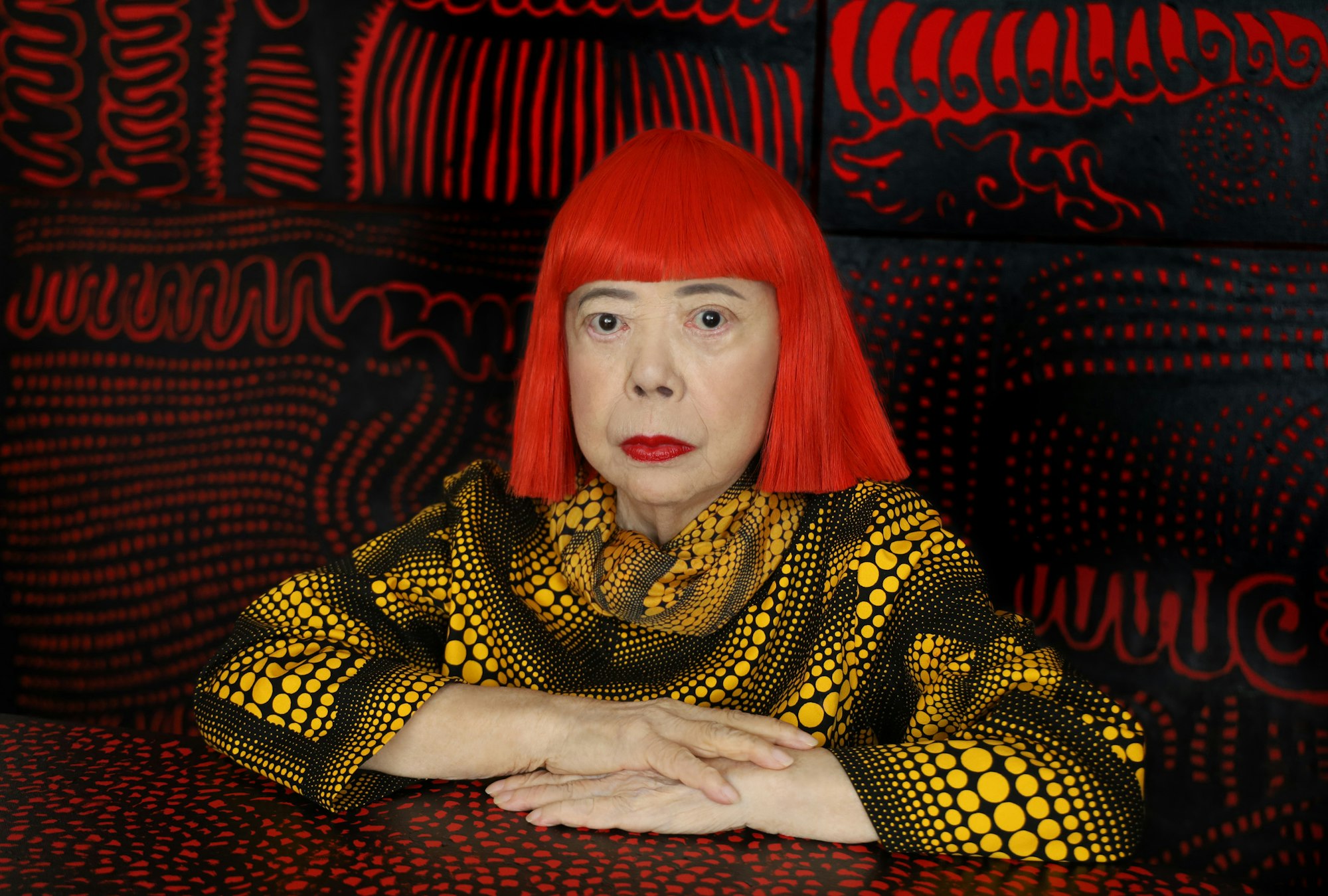A person with bright red bobbed hair and a yellow-spotted black top  in a red and black patterned space.