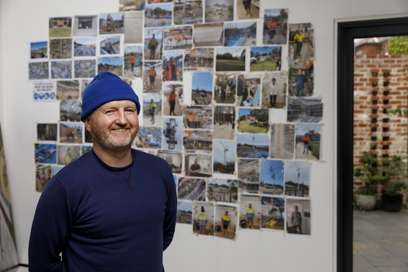 A person wearing a blue beanie stands in front of a wall holding a multitude of unframed photos.