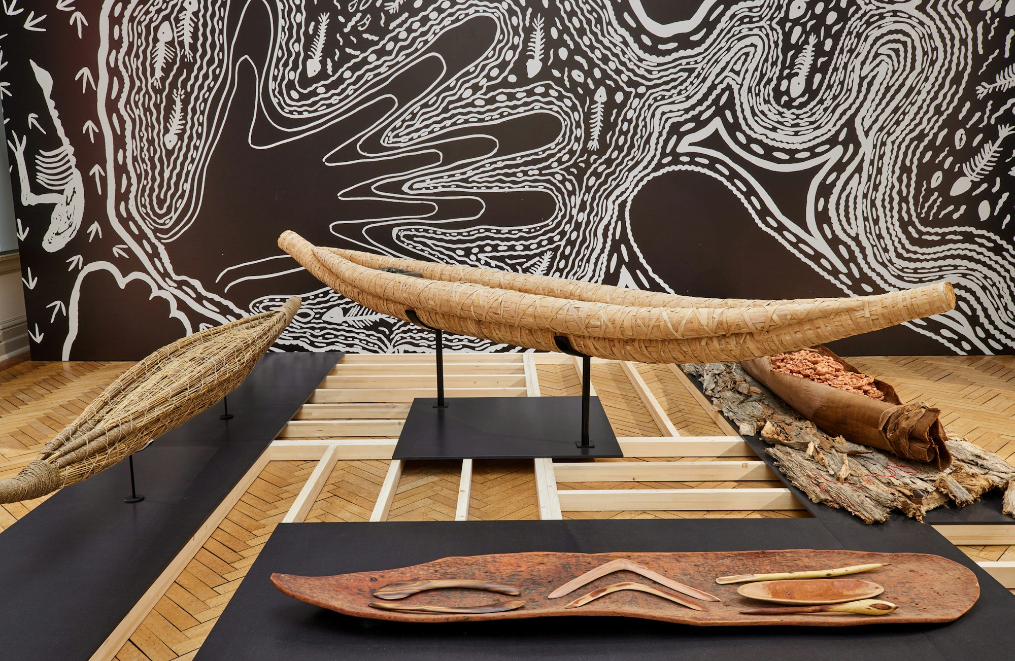 Bark canoes, boomerangs and other wooden objects in front of a black-and-white artwork featuring large hands.