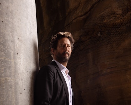 A person with a beard and short, wavy hair, dressed in a suit and open-necked shirt, stands against a concrete wall with their hands in their pockets.