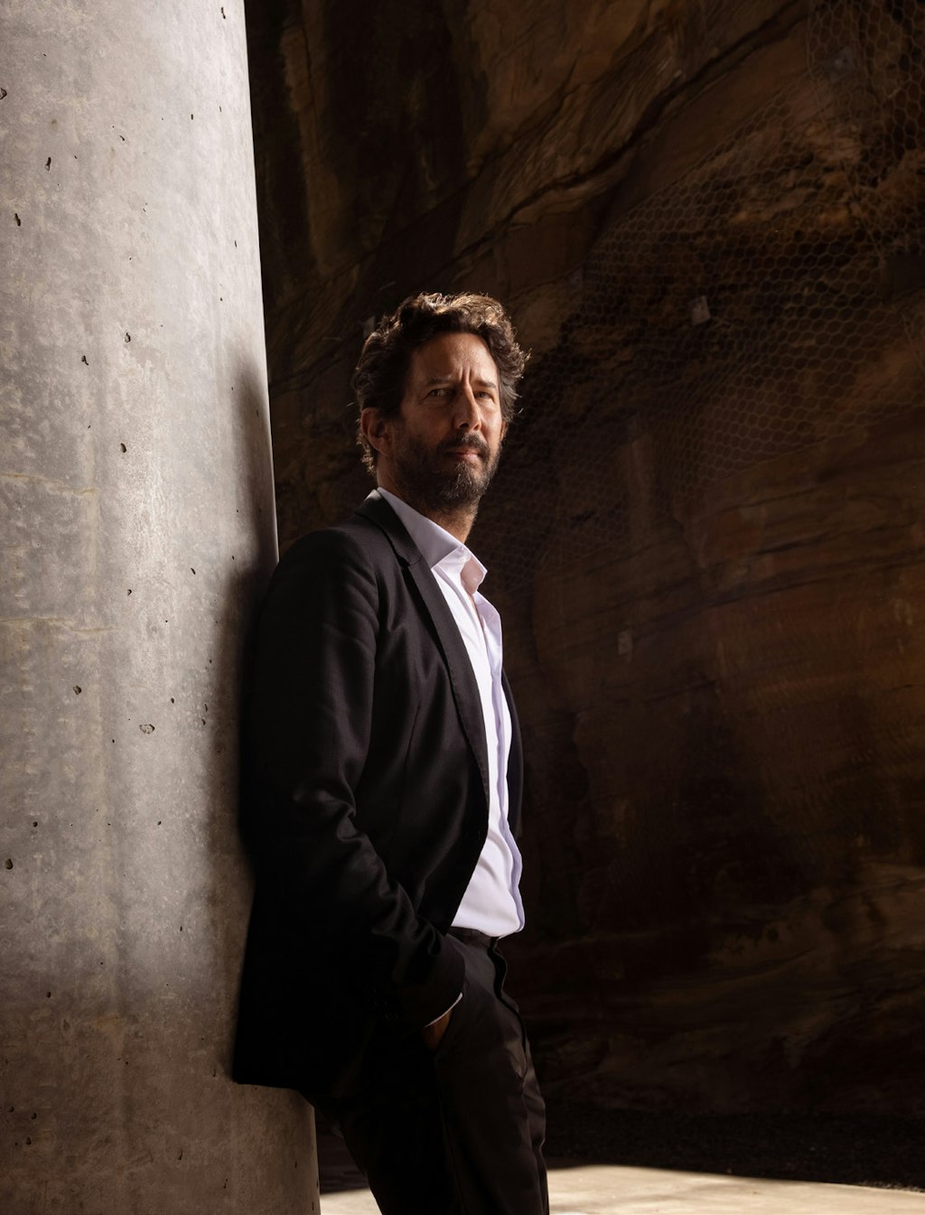 A person with a beard and short, wavy hair, dressed in a suit and open-necked shirt, stands against a concrete wall with their hands in their pockets.