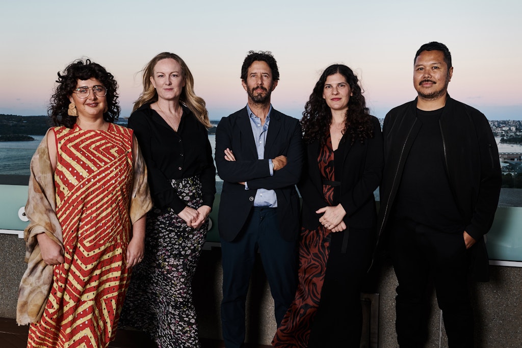 Five people of different genders and cultural backgrounds stand in a group with a view of water and land in the background.