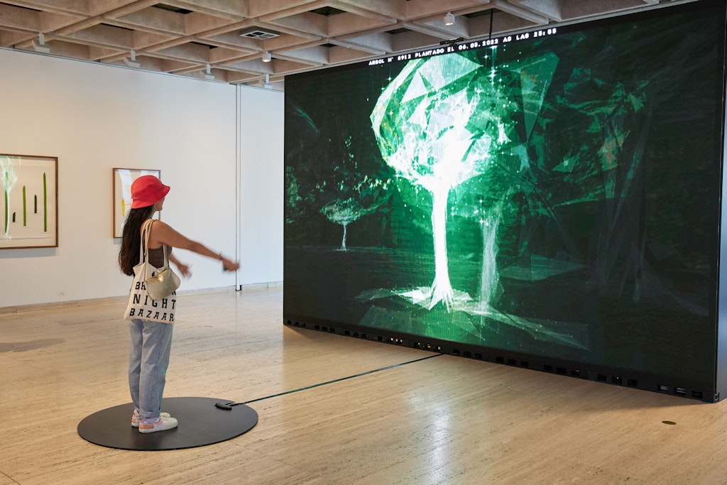 A person stands with their arms outstretched in front of them, facing a large screen with a green-and-white image of a tree.