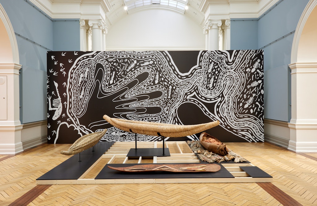 In an historic gallery space, several canoes and other objects are displayed in front of a large black-and-white artwork.