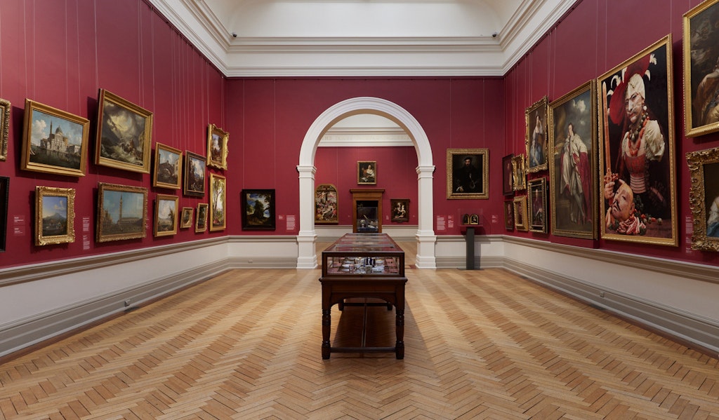 An historic gallery space with red walls hung with many gold-framed paintings. A glass-and-wood cabinet displaying ceramics sits in the middle of the wooden floor.