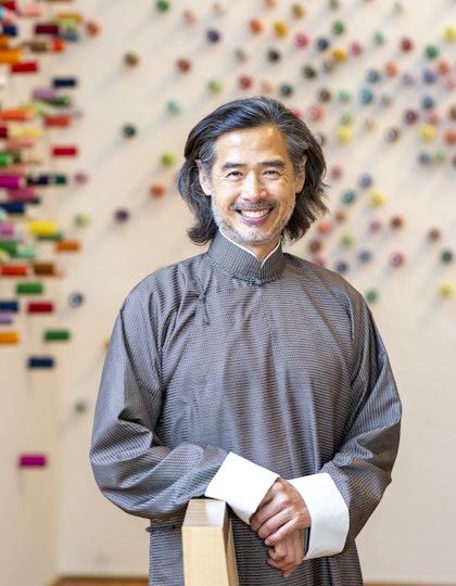A person with shoulder-length hair and a short beard, dressed in a grey robe with white cuffs.