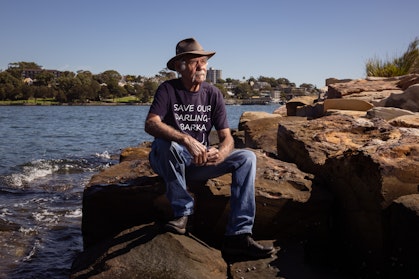 A person wearing jeans, t-shirt, boots and hat sits on rocks by the water.